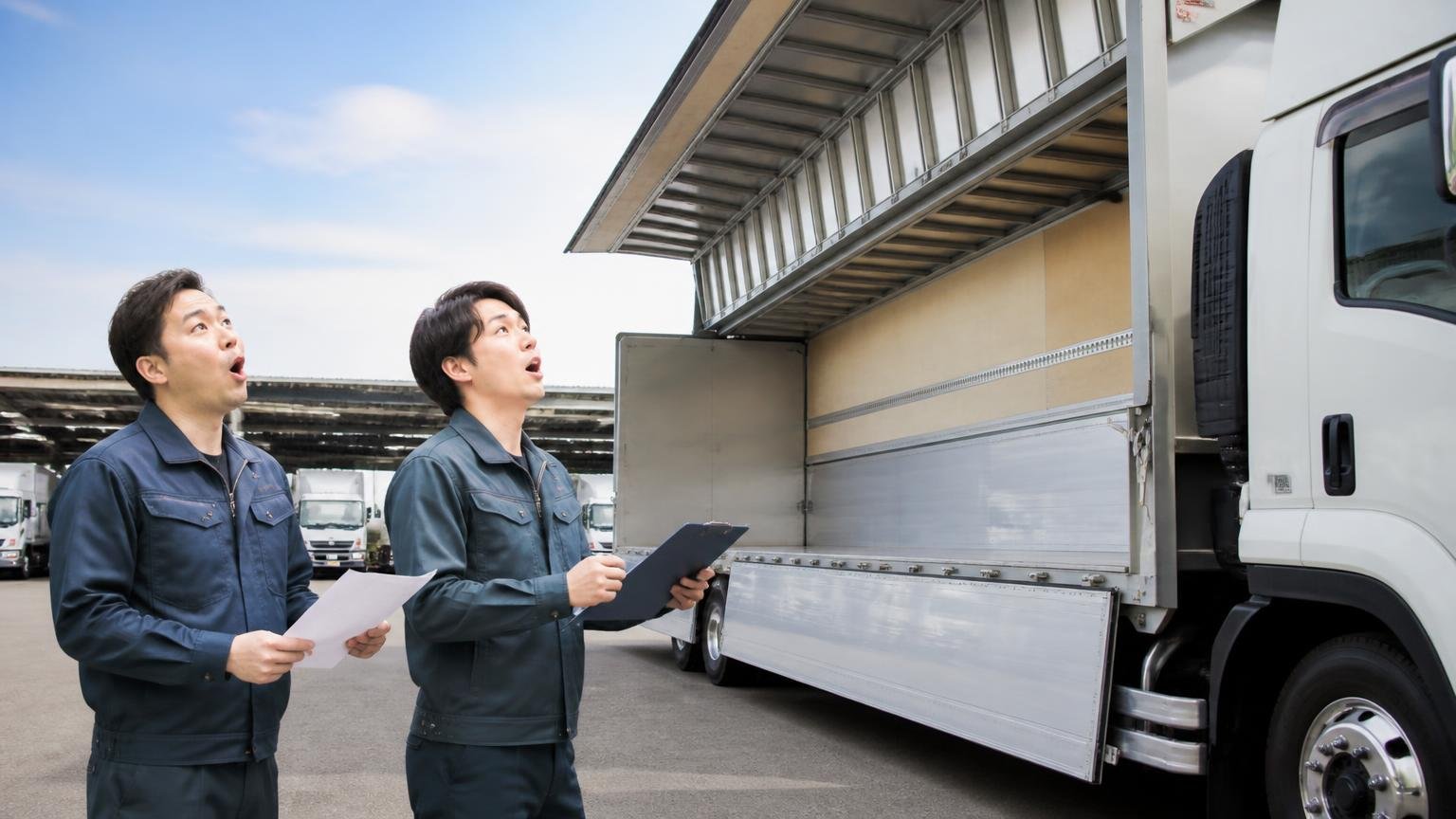 在庫が並ぶトラック展示場の一角。作業着の男性2人が資料を手にウイング車を見上げて驚いた表情