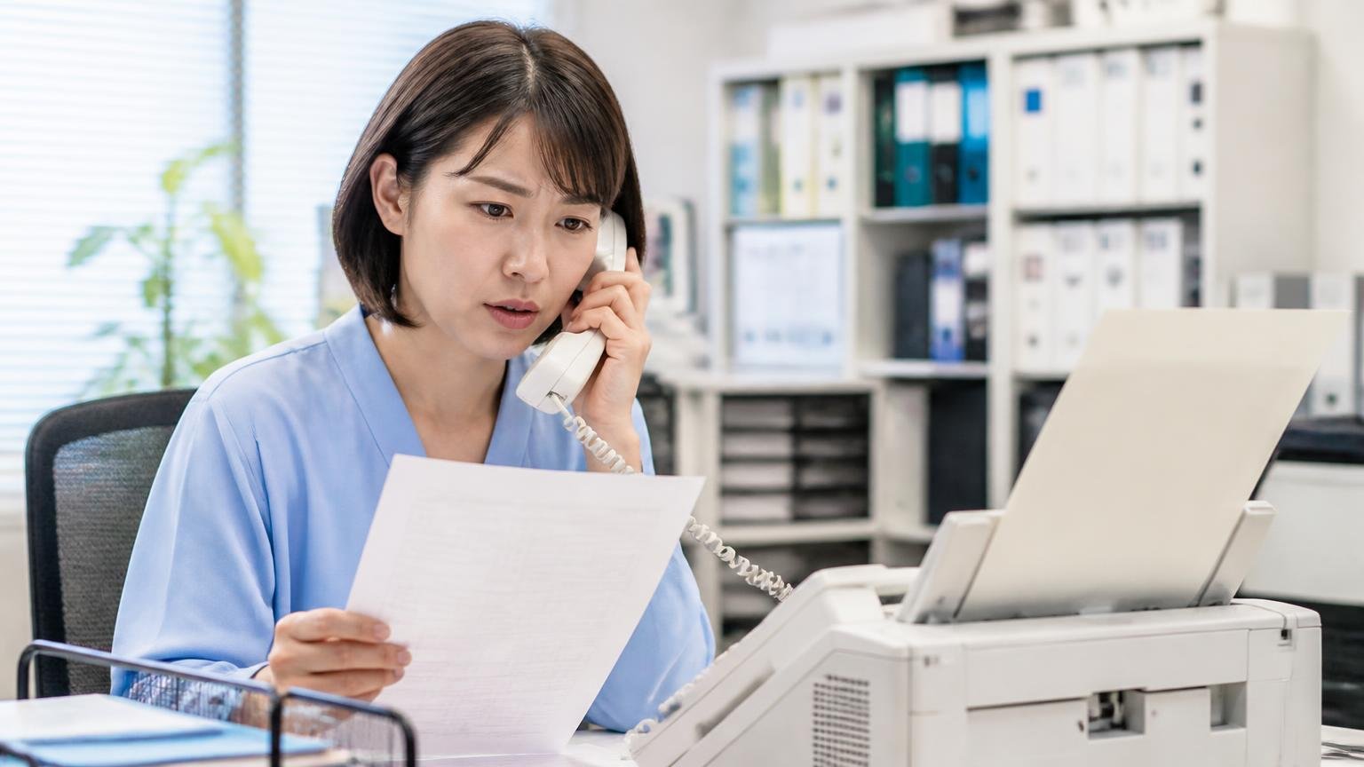 FAX停止の相談先を確認する場面。事務所の机で受信用紙を手に電話をかける事務担当1人が真剣な表情