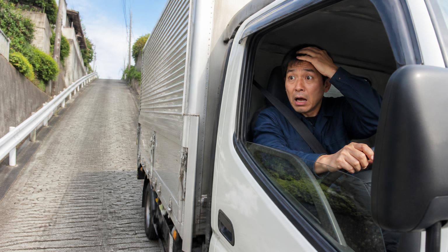 坂道で少し下がってから上り始める|急な上り坂の途中で停まったトラック運転席で焦る男性1人