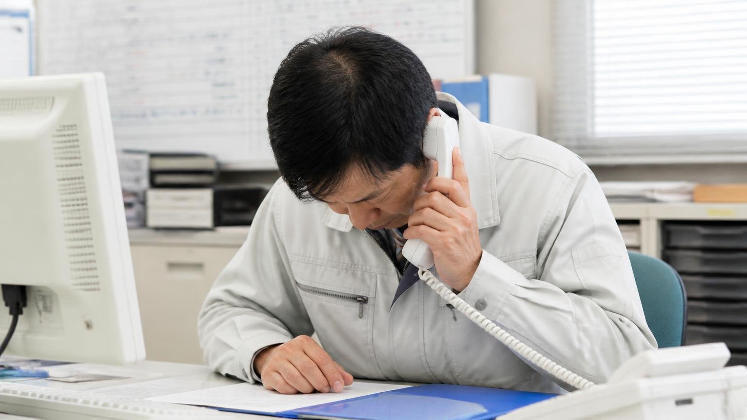 会社側の返信。営業所のパソコン前で受話器を持ち深く頭を下げる作業着の男性1人が真剣な表情