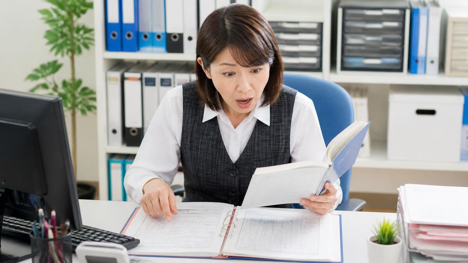 オイル交換時期が見直された 事務所で整備記録簿とメンテナンスノートを広げる机 事務担当1人 驚いた表情