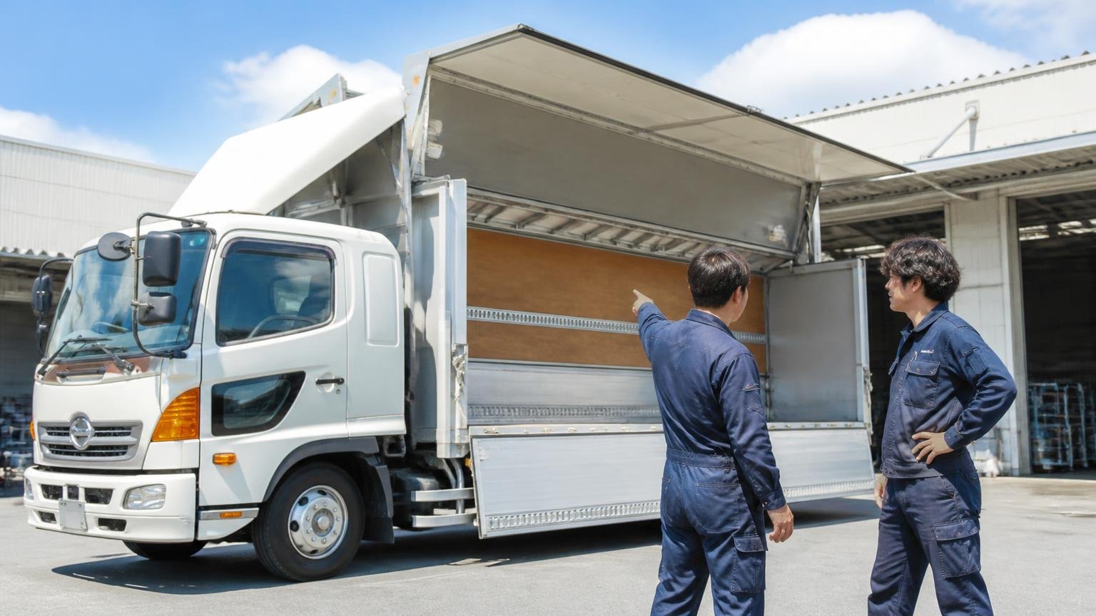 トラックセンターサンワが向いている人。営業所の車庫に停まった4tウイングの前。作業着の男性2人が荷台を指さして話す