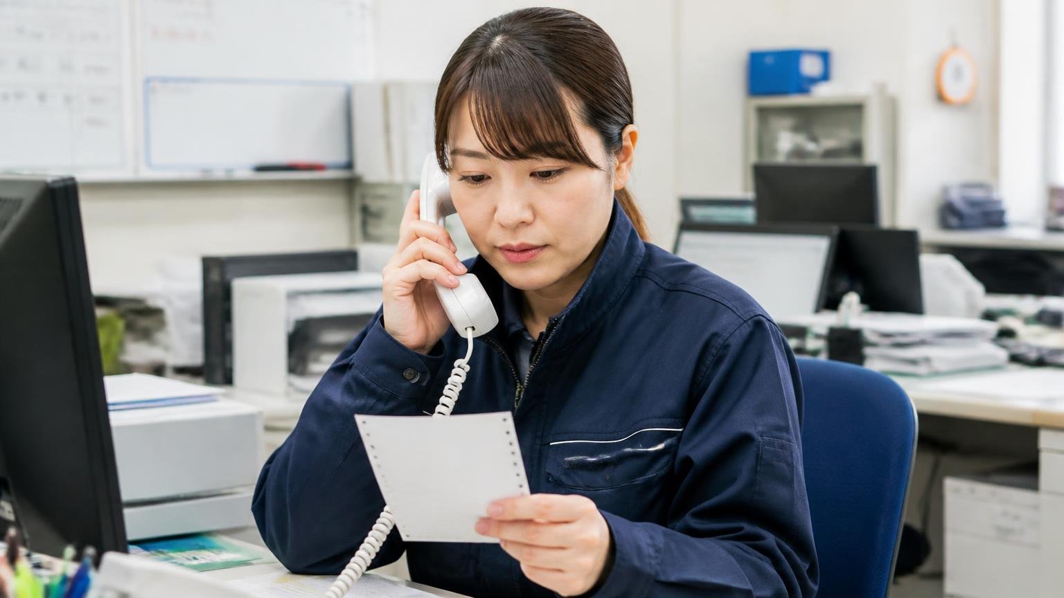 明日やることは連絡と査定の予約｜運送会社の事務所で受話器を持ちメモを見ながら話す30代の女性