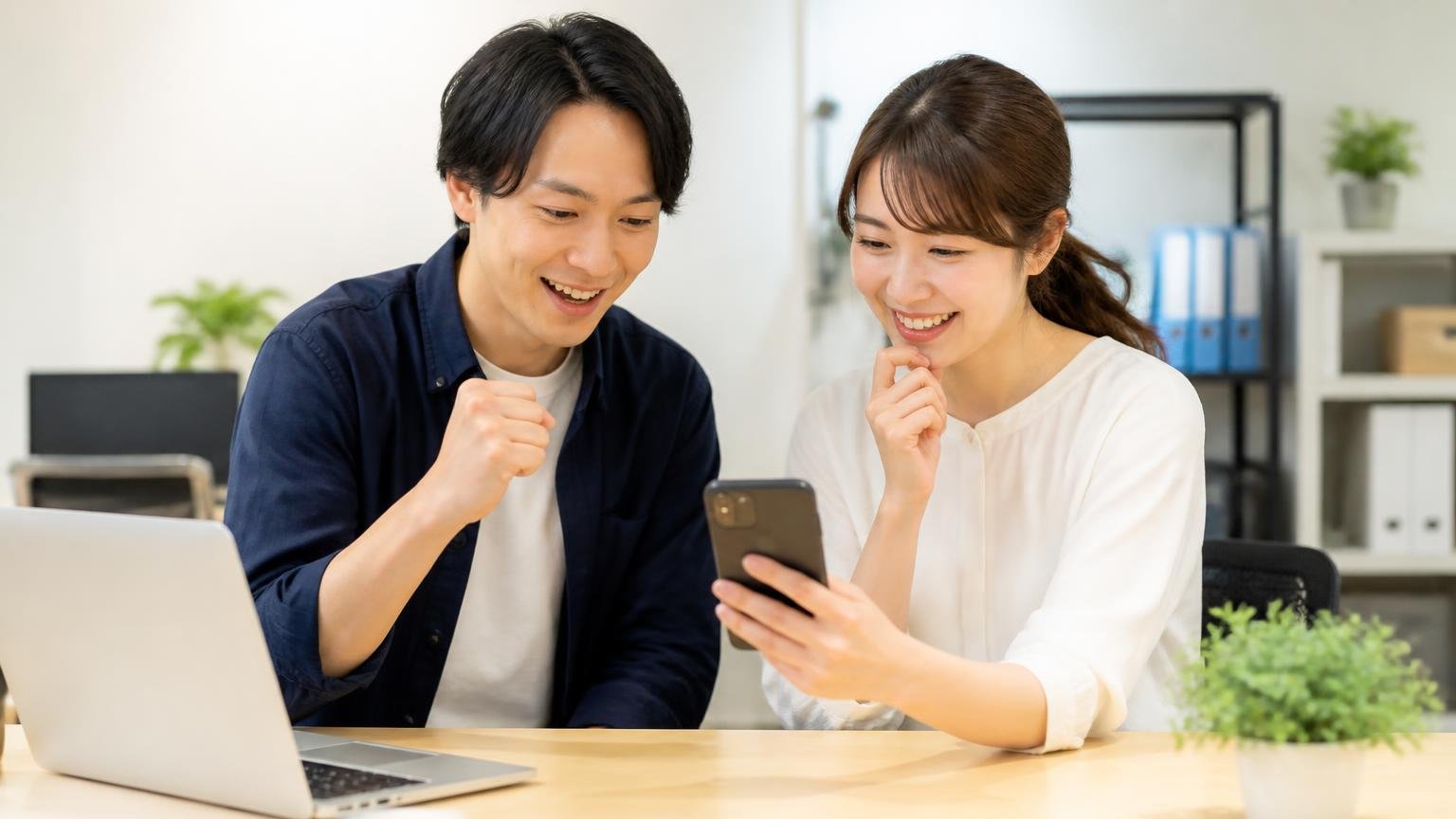 評判を読み終えたらあとは金額を横に並べるだけ｜事務所でスマホから無料査定を申し込む男女2人が前向きな表情