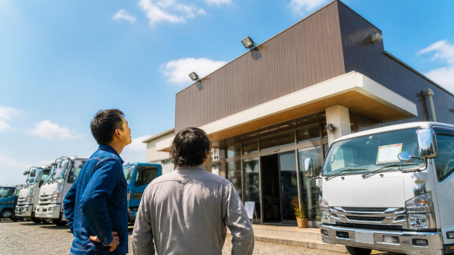 2001年設立、古物商の許可番号を公開している商用車の専門店、中古トラックが並ぶ屋外の展示場、店を見上げる男性2人