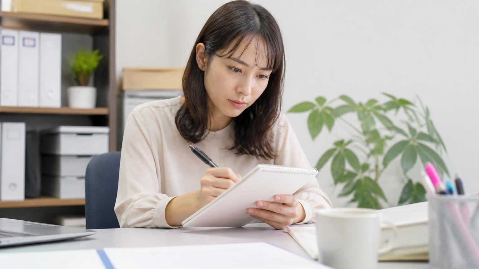 公式サイトに書かれていないことも確認材料になる|事務所の机でメモ帳に質問項目を書き出す女性1人が真剣な表情