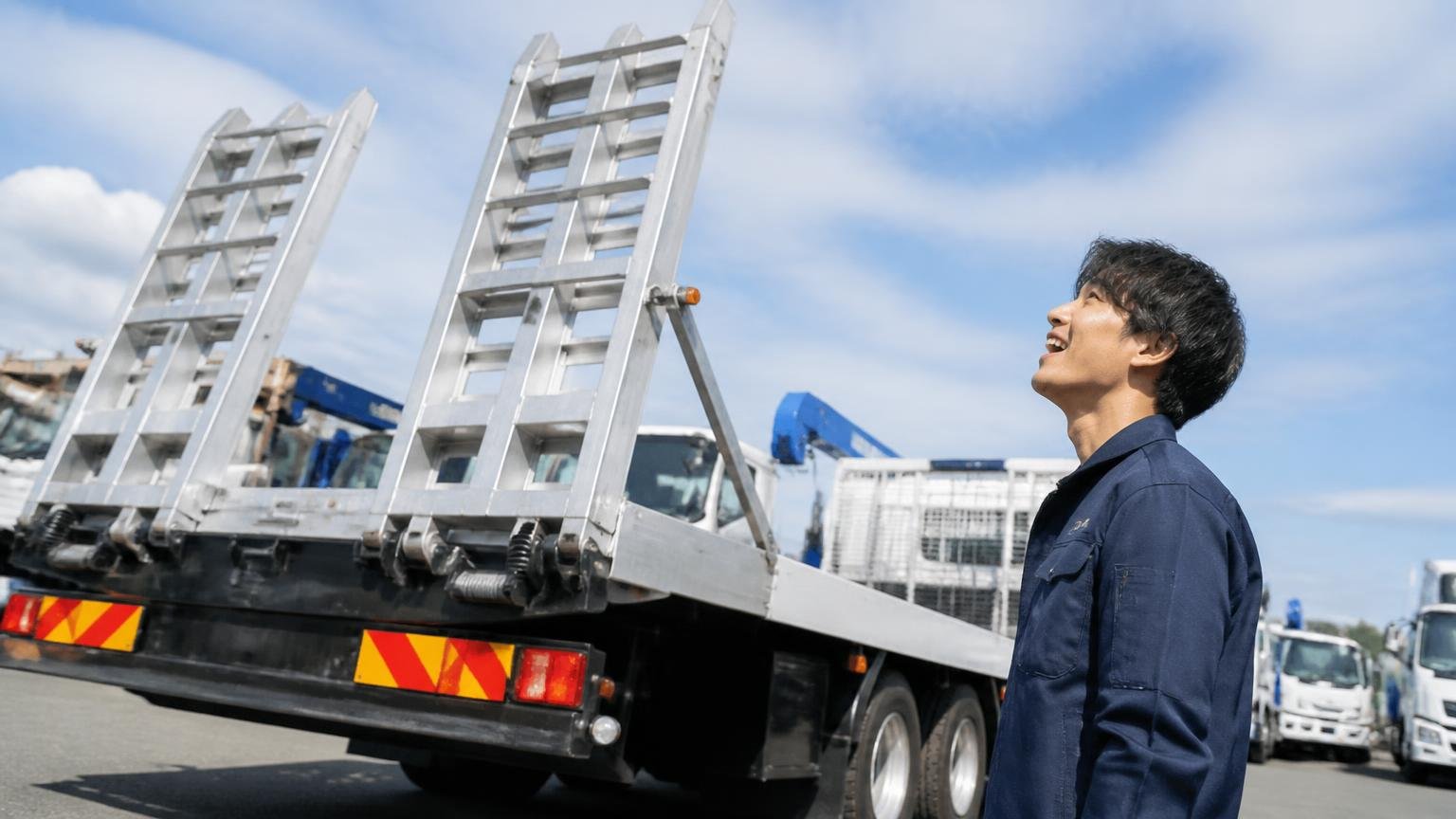 扱うのは重機運搬車・ダンプ・トレーラーなど"働く架装"が中心 屋外の中古トラック展示場で重機運搬車を見上げ目を輝かせる若いドライバー1人