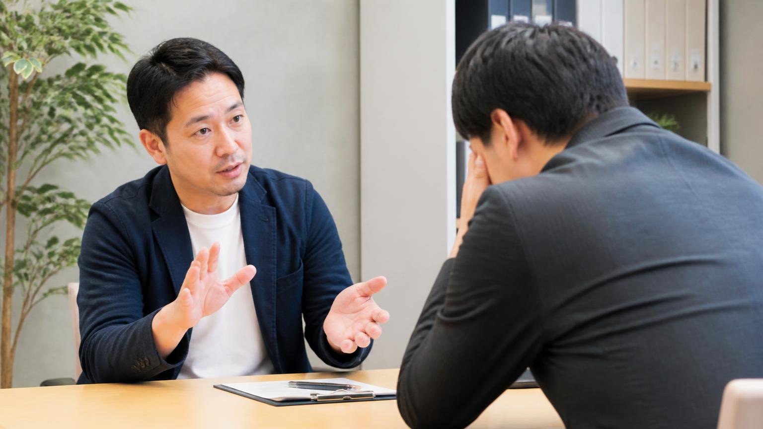 ★1を付けた人を責めてもあなたの査定額は変わらない／事務所の打ち合わせ席で向かい合う2人／片方が落ち着いた手振りで諭す
