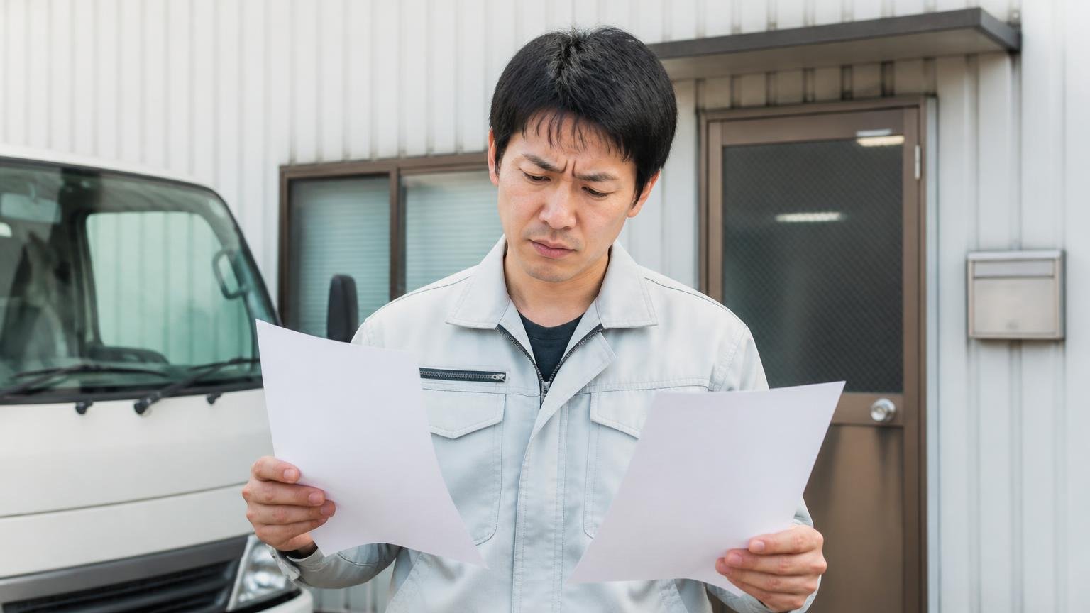 実在する古い会社と高く買う会社は別物、営業所の前で2枚の見積書を見比べる作業着の男性1人、眉をひそめる
