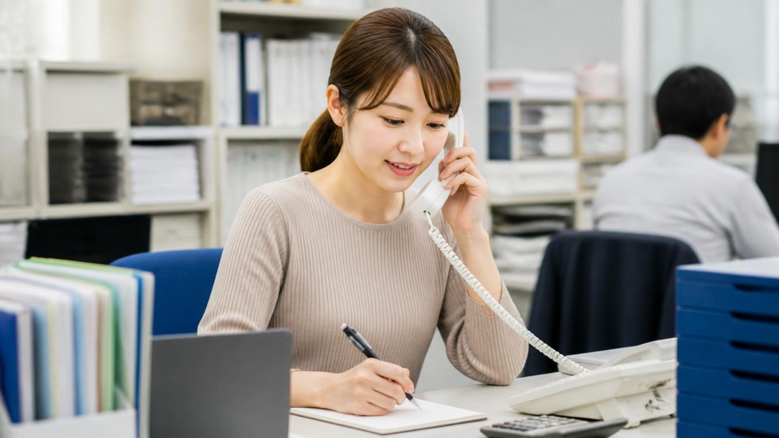 聞くべき3項目は査定料と対応エリアと書類代行｜営業所の事務所で電話をかけながらメモを取る30代の女性事務員