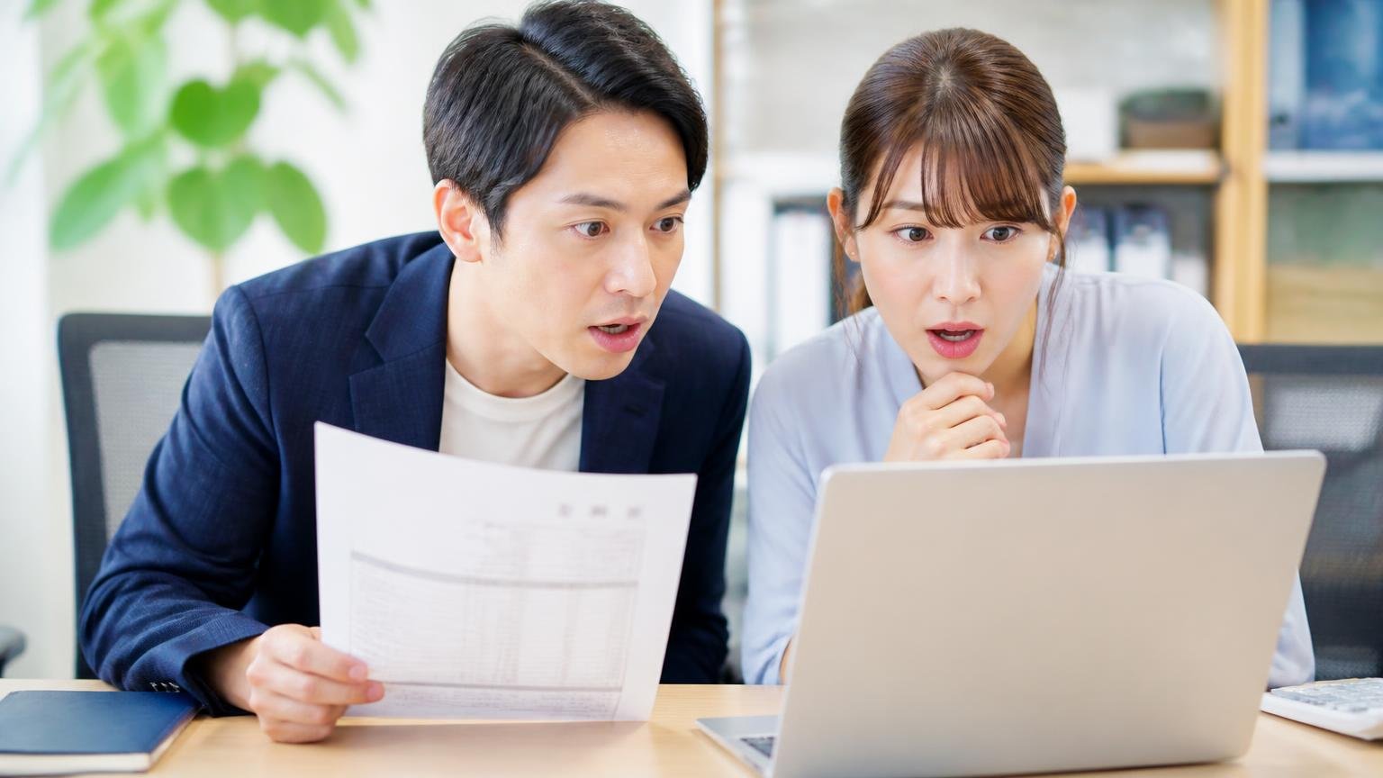 公式サイトで確認できる良い条件・事務所で見積書とパソコンを見比べる男女2人が身を乗り出す