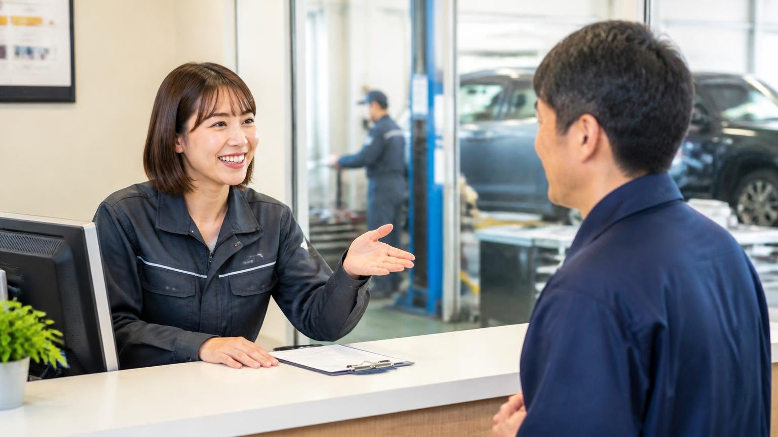 来客を温かく迎えてくれる声。整備工場の受付カウンターで来客を迎える場面。女性スタッフと男性客が笑顔で会話