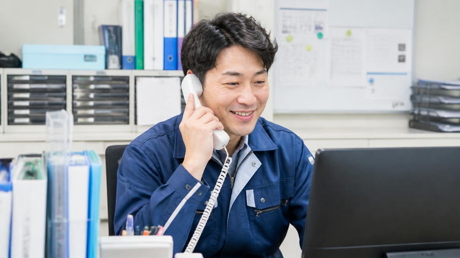 「顔の見えない取引でも心遣いが伝わる」遠方とのやり取りがスムーズという声、事務所で電話しながらメール画面を確認しほっとした表情の運送会社の男性1人