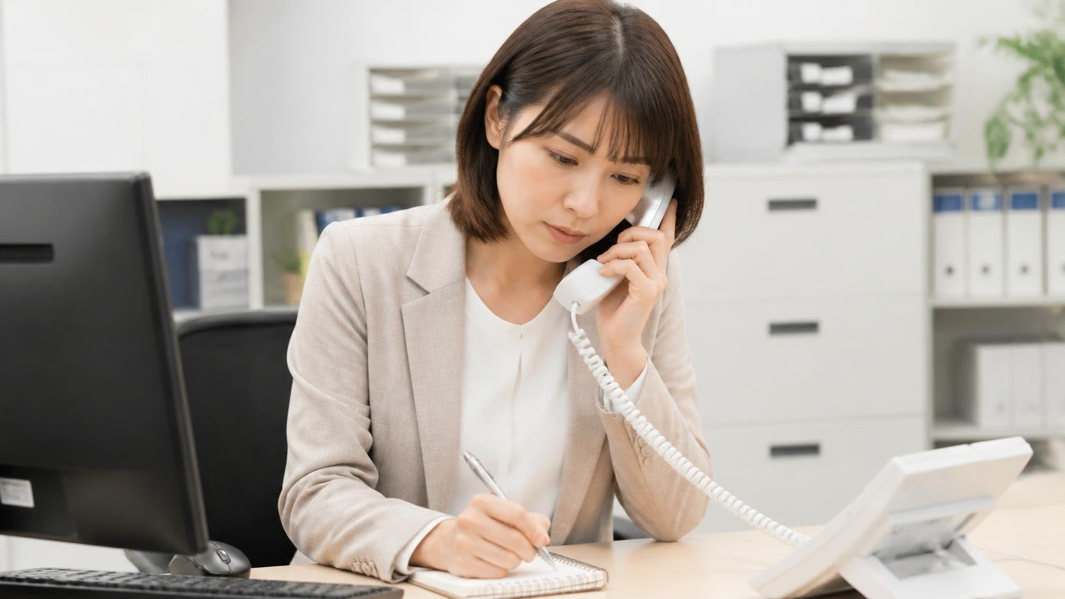 出張査定や査定料の記載がなく電話で最初に確認する項目、事務所で受話器を持ちメモ帳に確認項目を書く30代女性1人、真剣