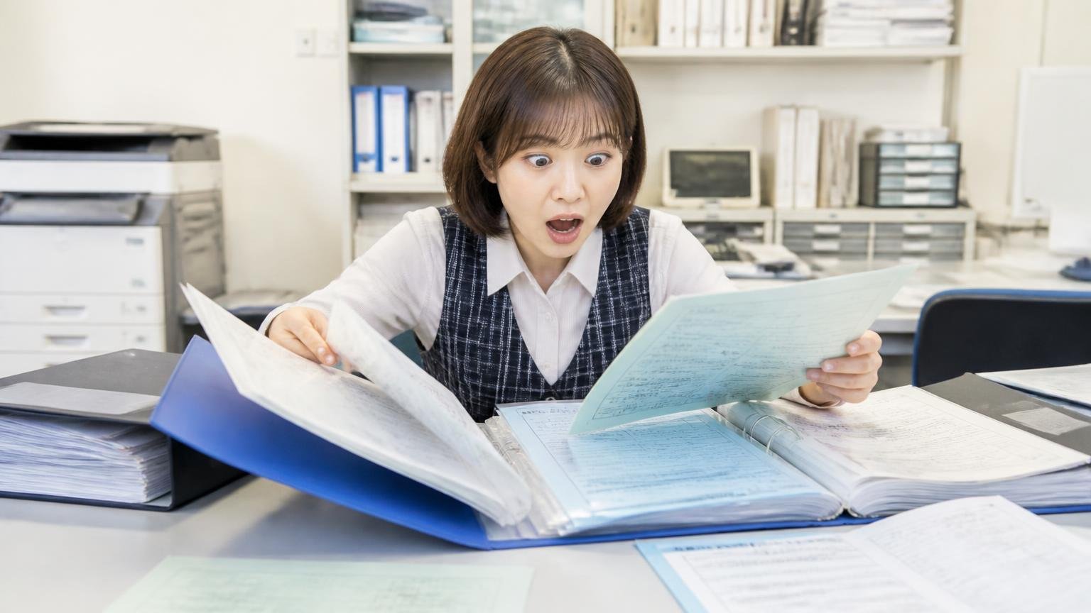 車検証・整備記録簿・架装の書類をそろえる、事務所の机にトラックの書類ファイルを広げて探す女性事務員が目を丸くする場面