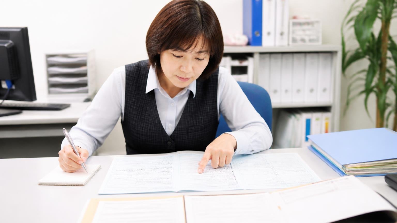 査定を頼む前に用意しておくもの。事務所の机に車検証と書類を広げた女性事務員1人が指さし確認しながらメモを取る
