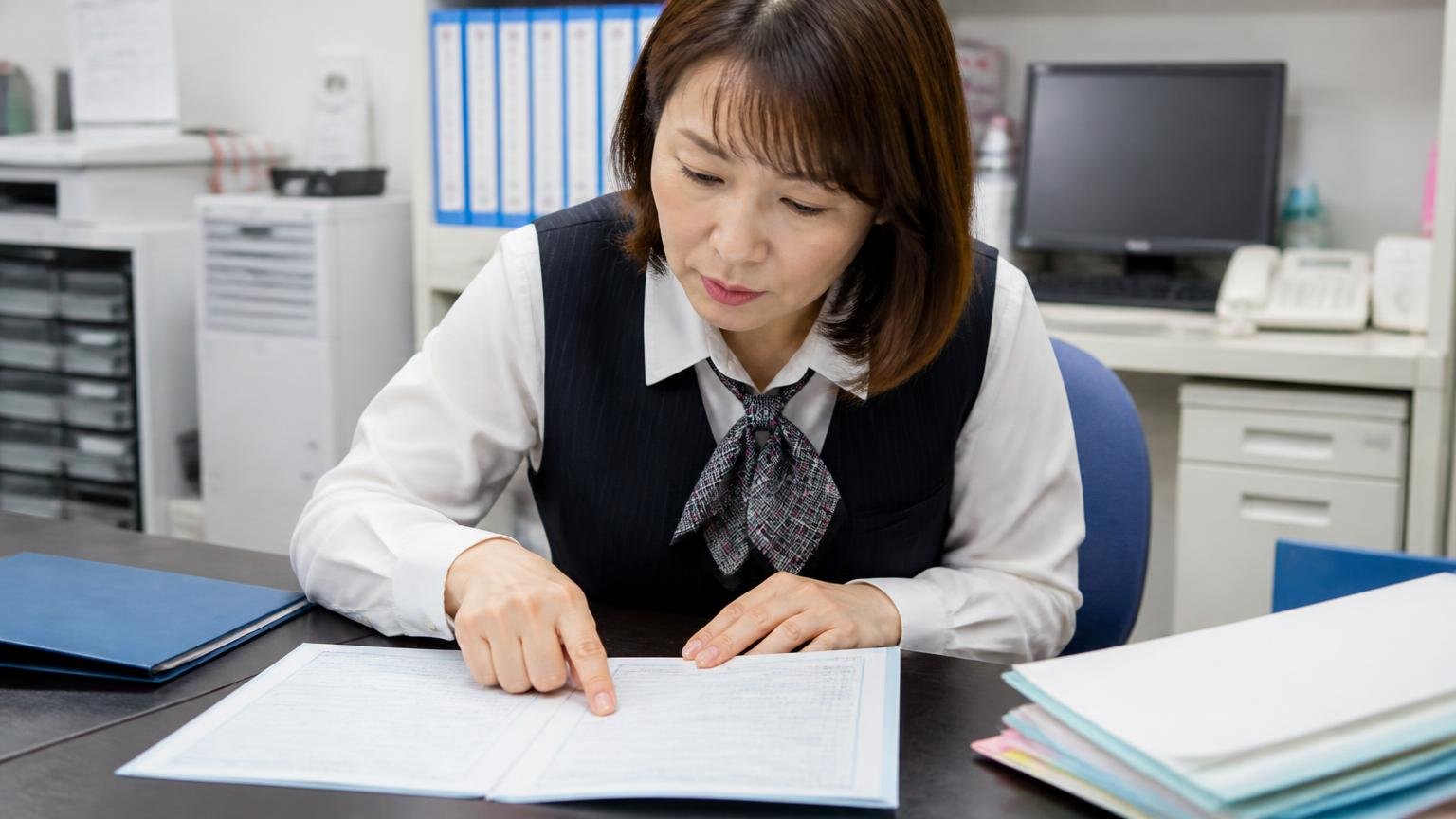 申込前にそろえる書類。運送会社の事務所の机で車検証を広げ所有者欄を指でなぞる事務服の女性1人が真剣な表情