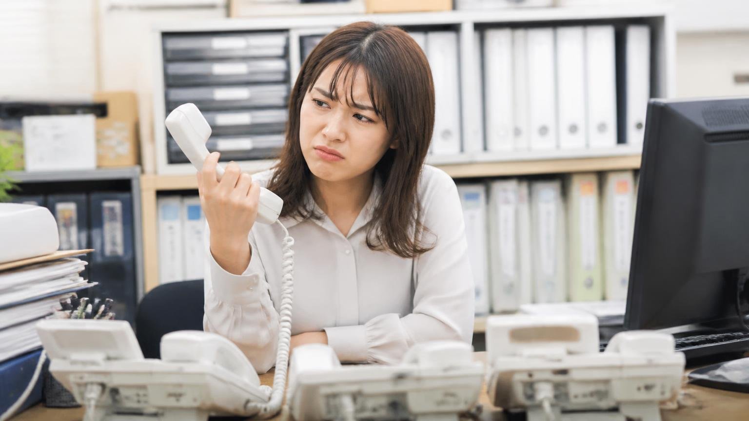 営業電話が多いという不満は査定額とは別の話。事務所で鳴り続ける電話を前に女性1人がうんざりした表情で受話器を見る