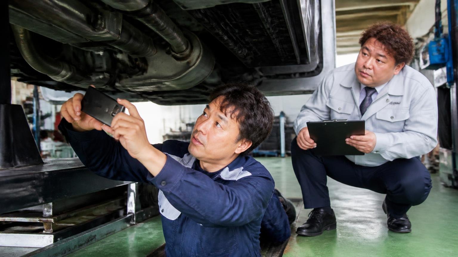 サビや不具合は先に伝える 車庫でトラックの下回りをスマホで撮影する運転手とそれを見守る査定員の2人