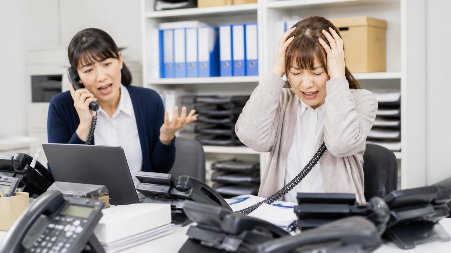 電話がしつこい今でも電話が来るという悪い口コミ、トラック買取の営業電話が鳴り続ける運送会社の事務所で頭を抱える女性事務員と同僚の2人