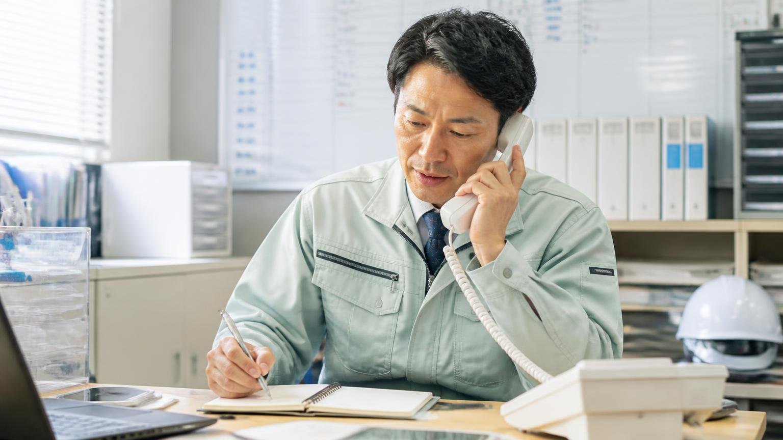 売却までの流れを確認する場面。営業所で電話を手に取りながらメモを取る作業着姿の男性1人