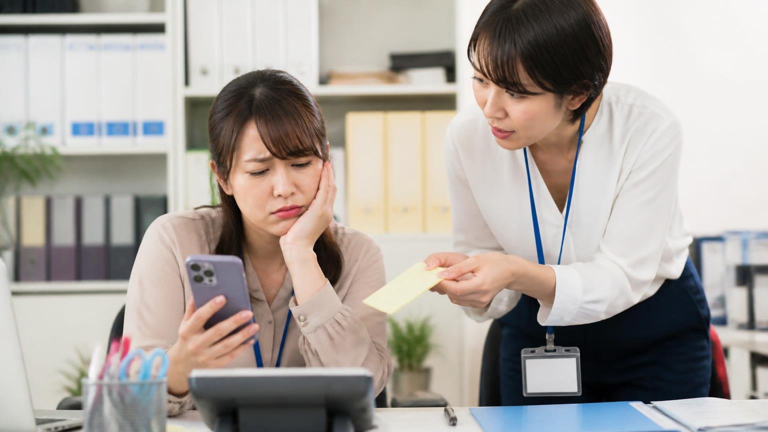営業電話が不安なら、連絡方法と社数を自分で決める 事務所でスマホの着信画面を見てため息をつく女性事務員と、メモを差し出す同僚