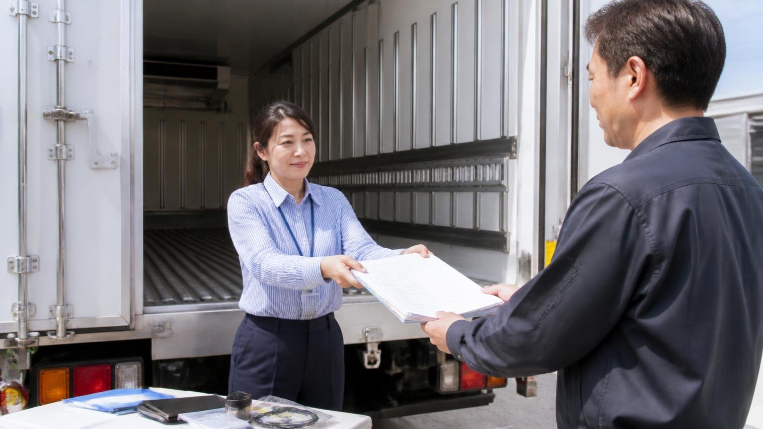 荷台や架装の不具合は隠さず伝え整備記録や付属品をそろえておく 冷凍車の荷台の前で整備記録簿を査定士に手渡す女性事務員