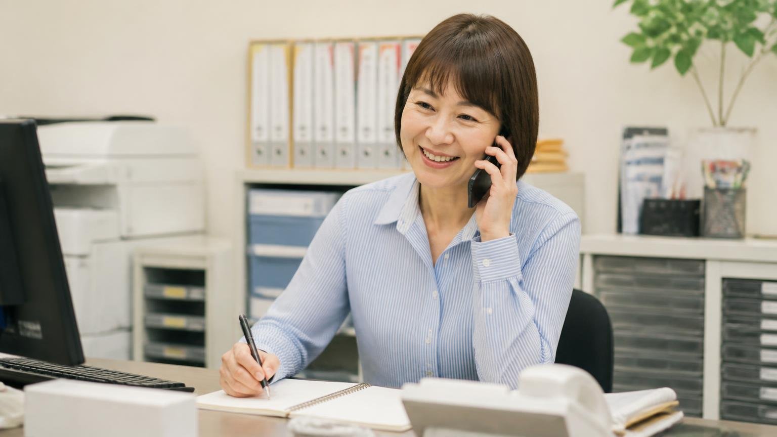 ワイズトラックを含めた2〜3社に無料査定を申し込む、事務所でスマホを耳に当てる場面、事務員の女性1人、メモを取りながら明るく話す