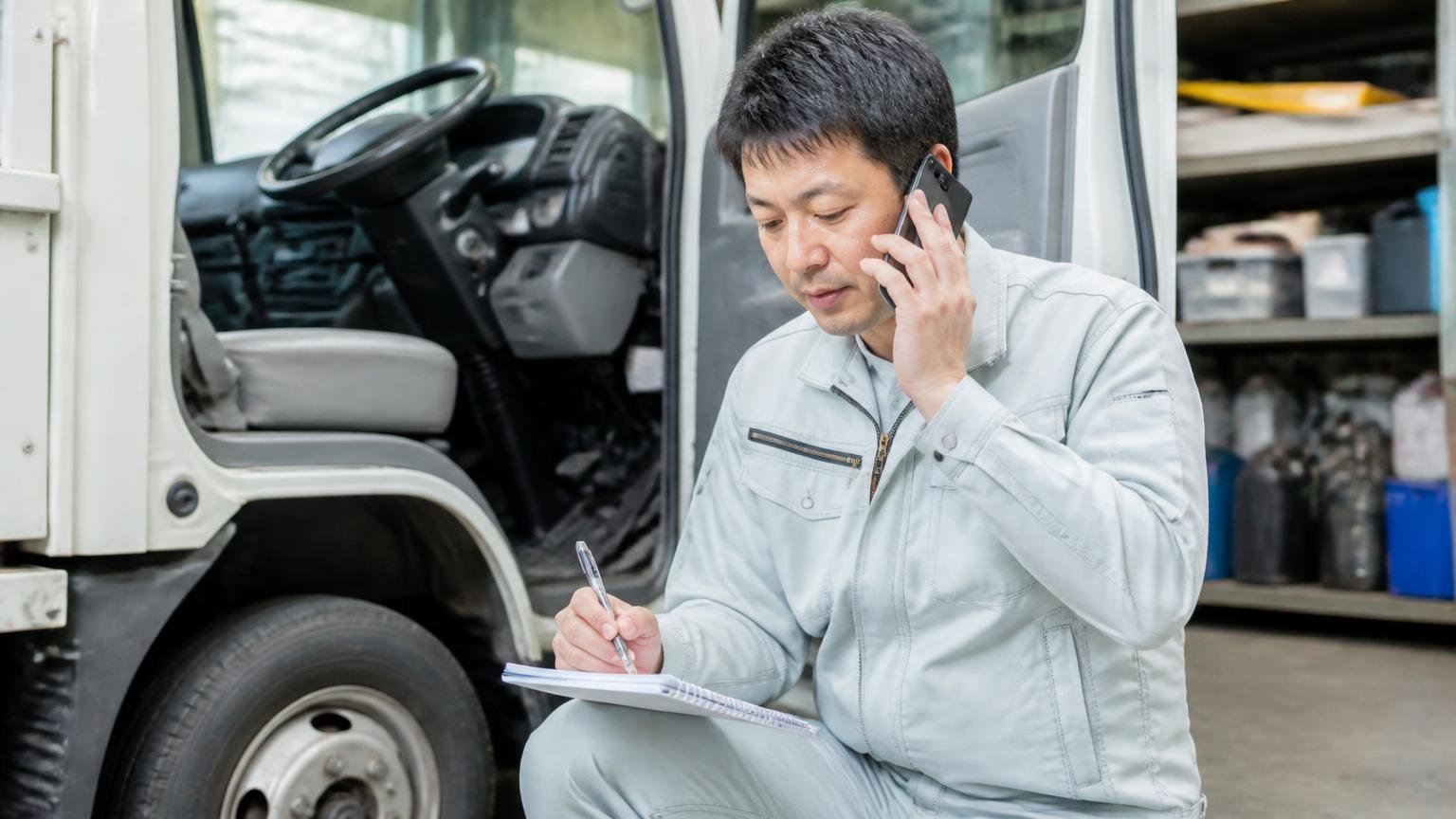 電話で必ず聞く5項目 車庫の運転席横でスマホを耳に当ててメモを取る作業着の男性1人