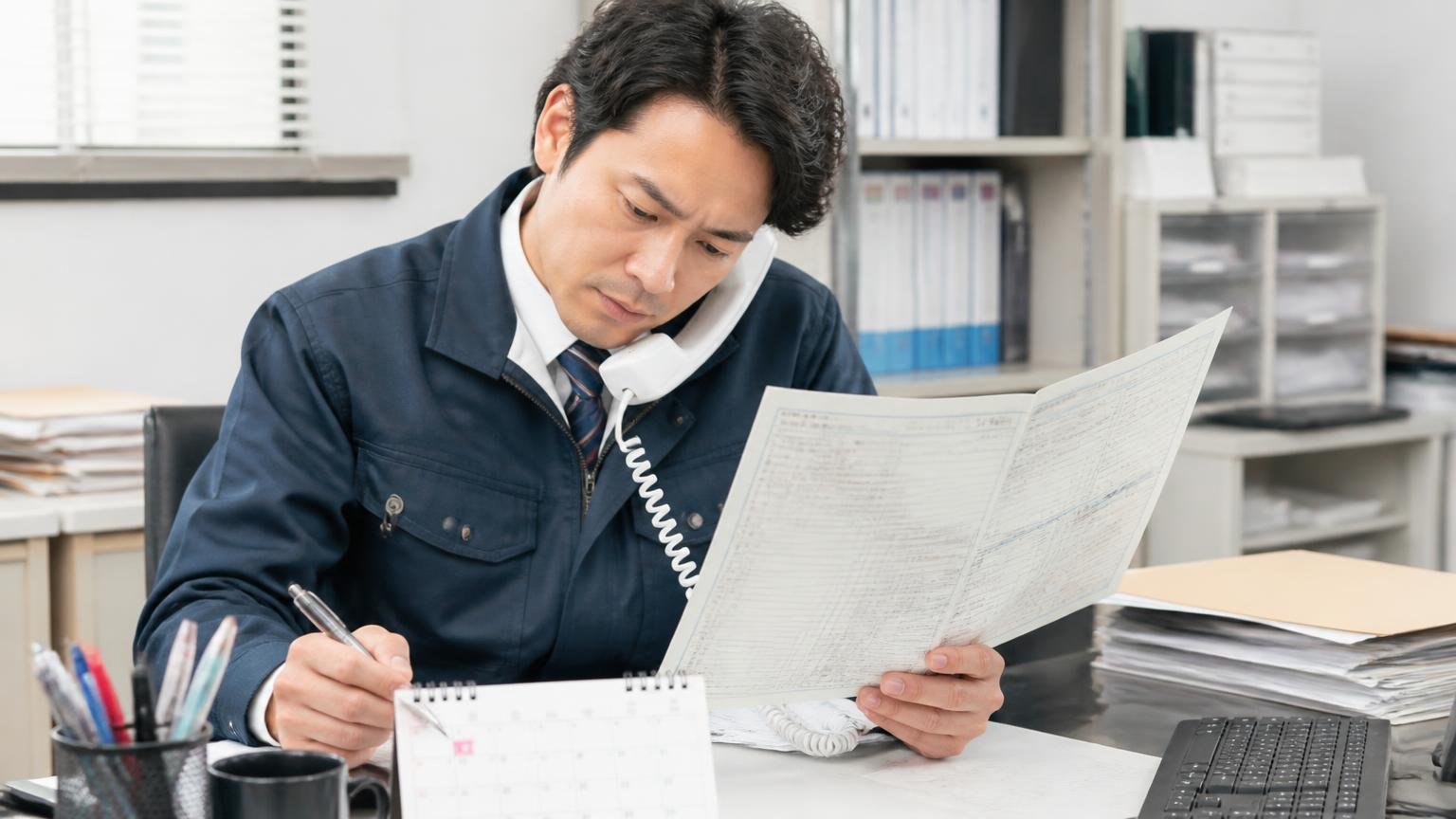 名義変更はいつまでに完了するのか確認する。事務所で車検証を広げた男性1人が電話しながらカレンダーに書き込む