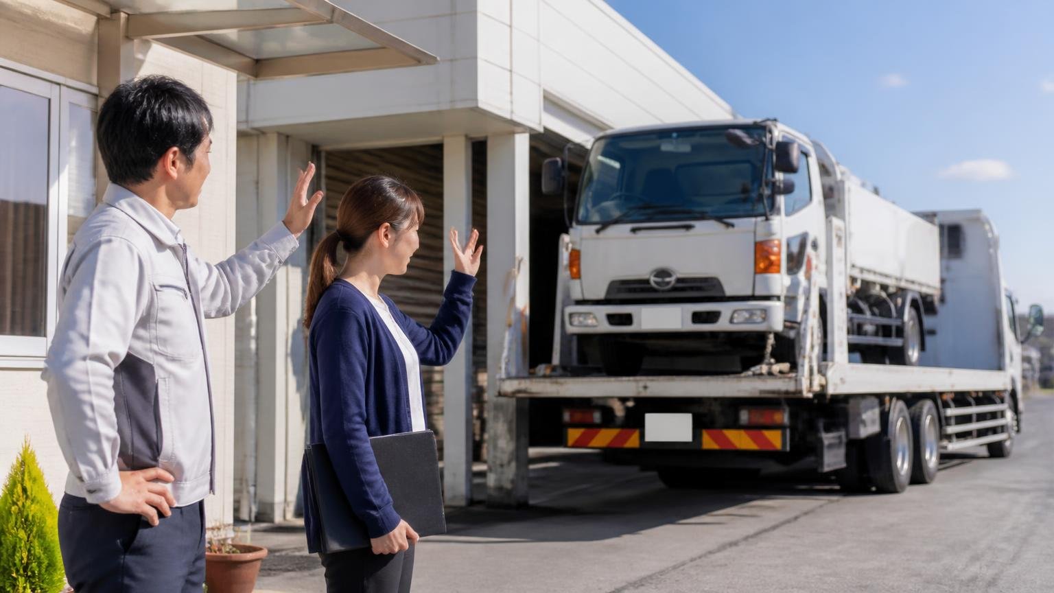 金額を横に並べる2社目をまだ決めていない人へ 営業所の前で積車に載せられるトラックを見送る場面 男性1人と女性1人 見送る穏やかな表情