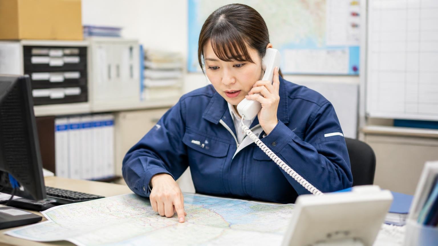 自分の地域まで出張・引取りに来てくれるかを確かめる 営業所の事務所で地図を広げ、電話で出張エリアを確認する運送会社の女性事務員1人