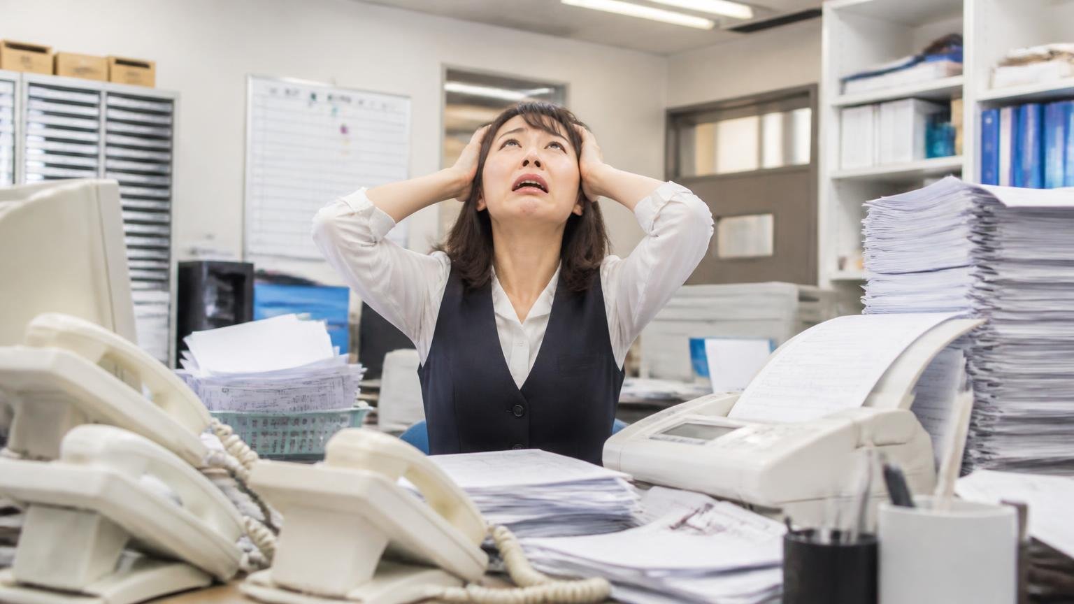 連絡手段や時間帯をこちらで指定できるか 運送会社の事務所 鳴り続ける電話とFAXの山を前に事務担当の女性が頭を抱えて天井を見上げる