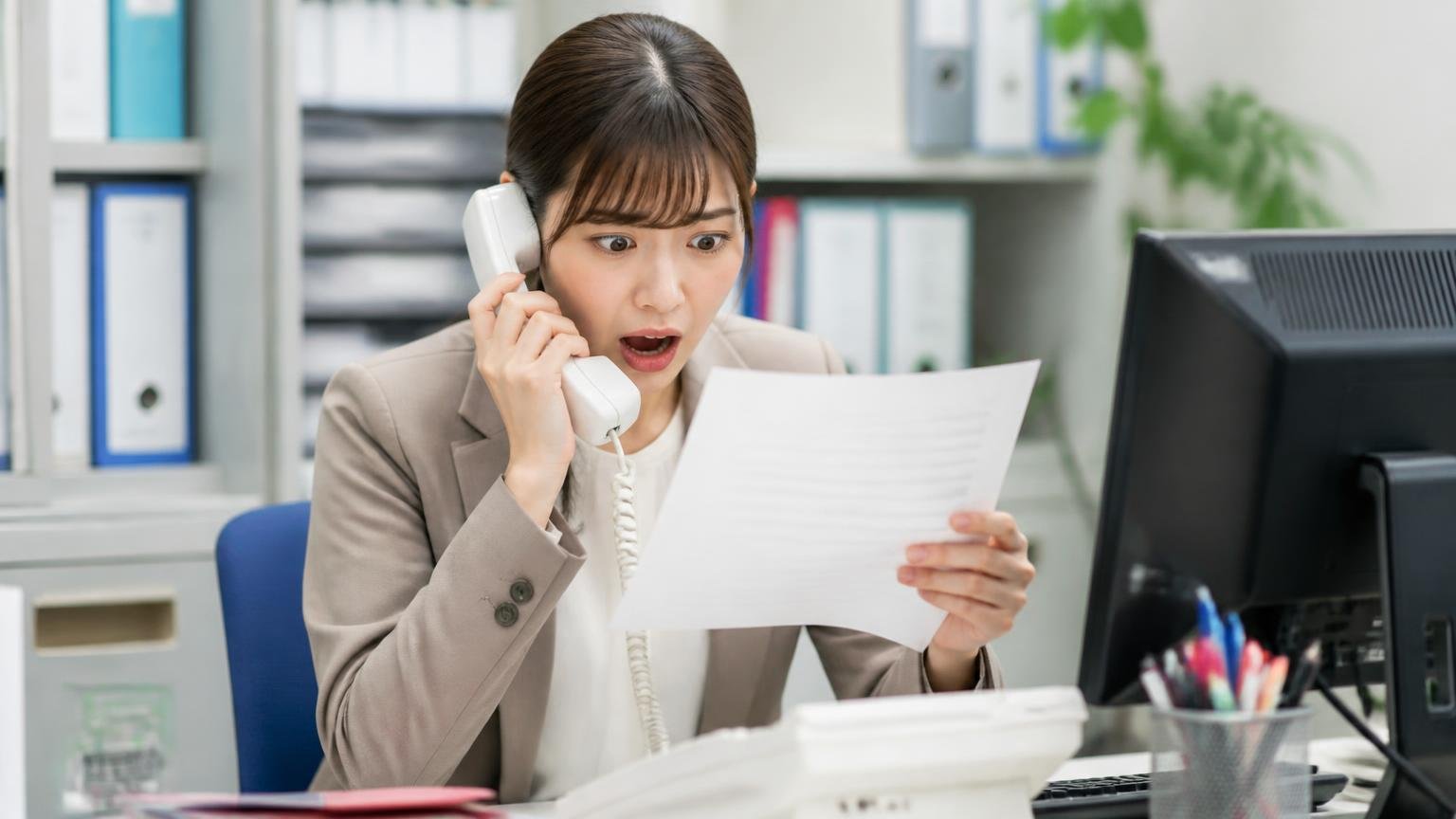 キャンセル料と代金の支払い条件を書面で確かめる 事務所の電話口 受話器を手に書類を見て驚き目を見開く若い女性