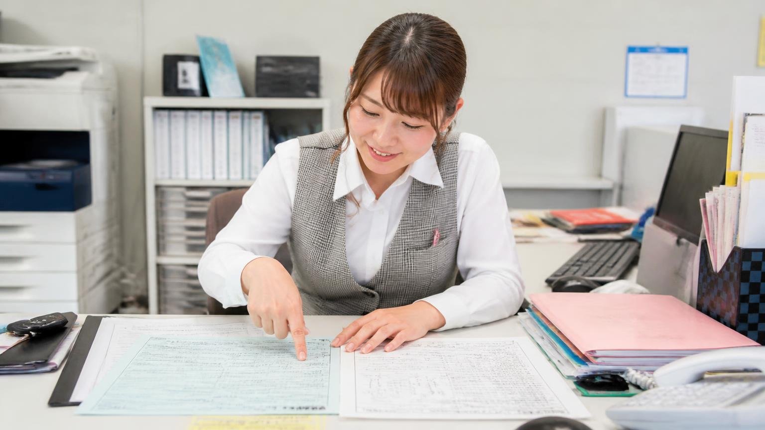 書類・付属品・整備記録をそろえ架装や故障箇所は自分から伝える|事務所の机に車検証と整備記録簿を並べて指差し確認する女性事務員1人