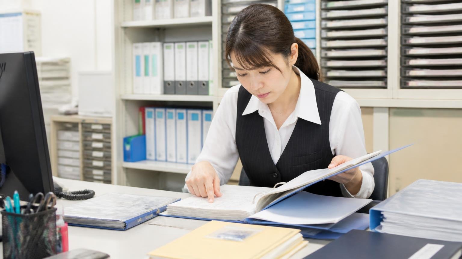 整備の記録・書類・付属品をそろえておく 事務所の棚の前 車検証や整備記録簿のファイルを机に並べて確認する女性事務員